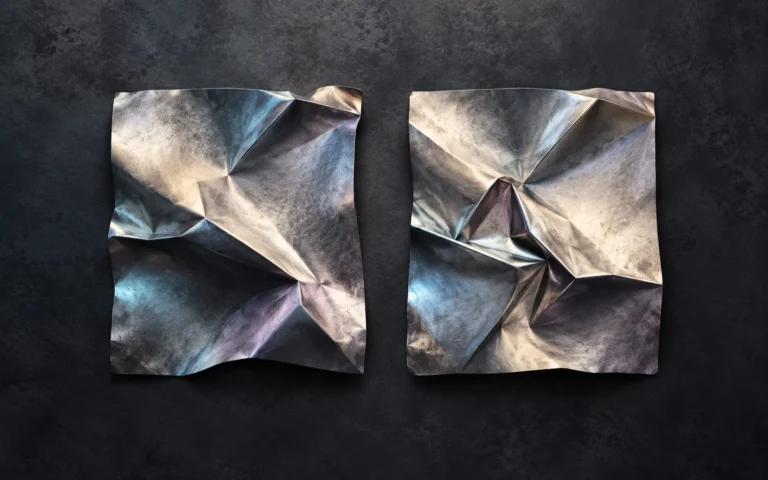 Two folded metal forms sit side by side, one rougher and one more polished.